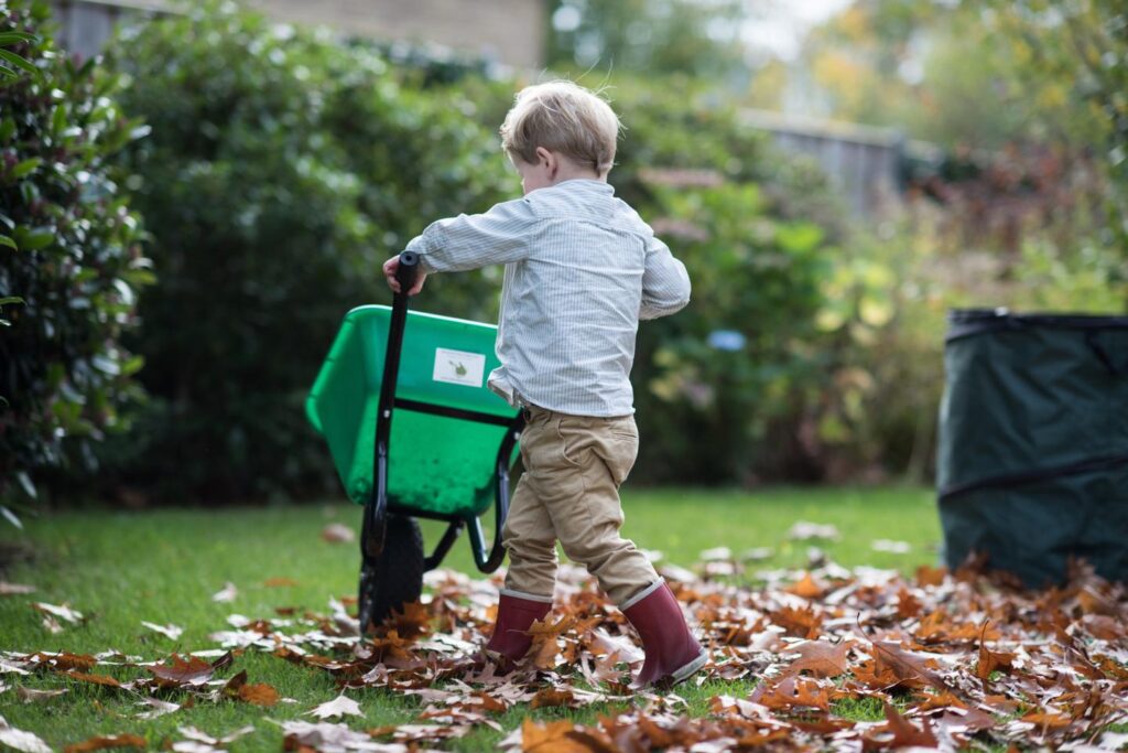 Jongen met rode laarzen en kruiwagen op gras met bladeren