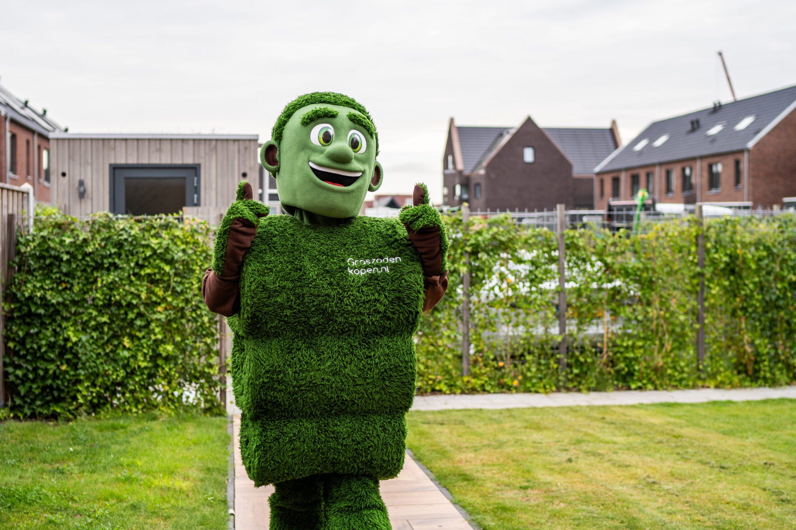 Grasmattie, de mascotte van Graszodenkopen.be, in een tuin met een vrolijke duim omhoog